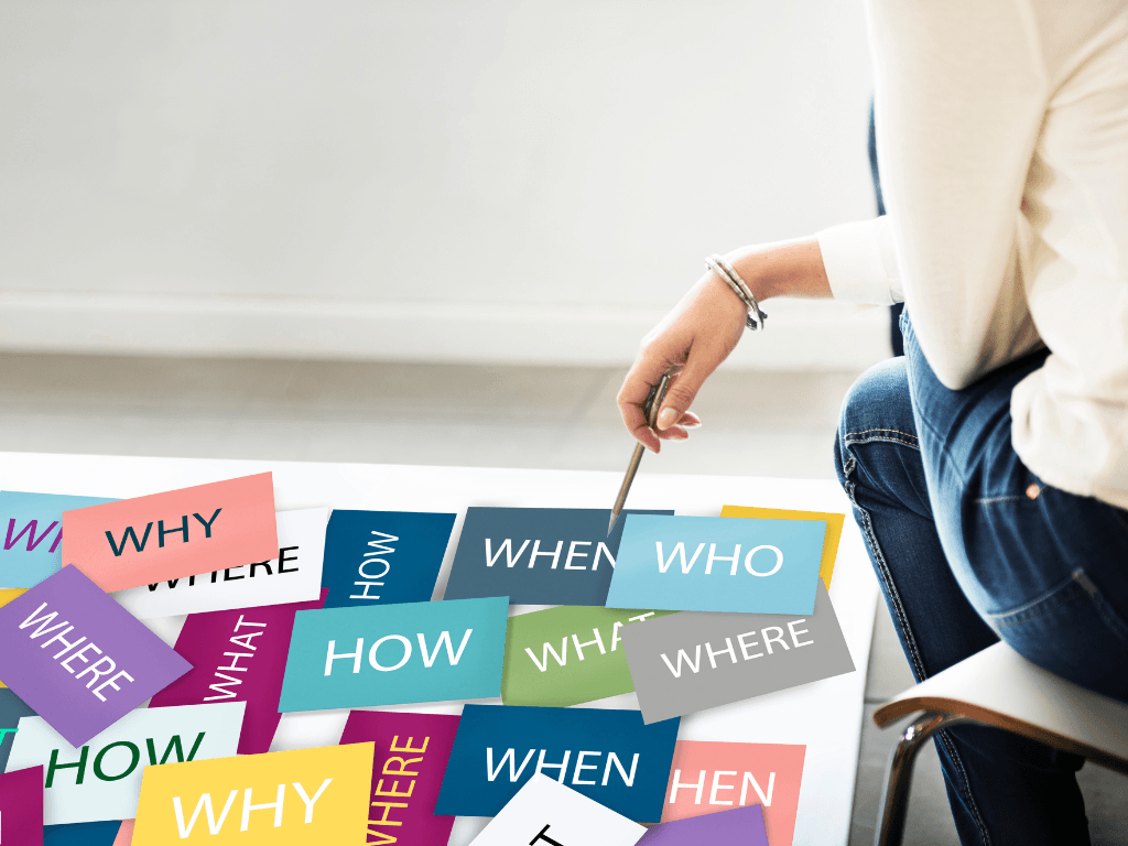 A hand holding a pen pointing towards a board filled with colourful keycard questions like: who, what, when, where, why and how