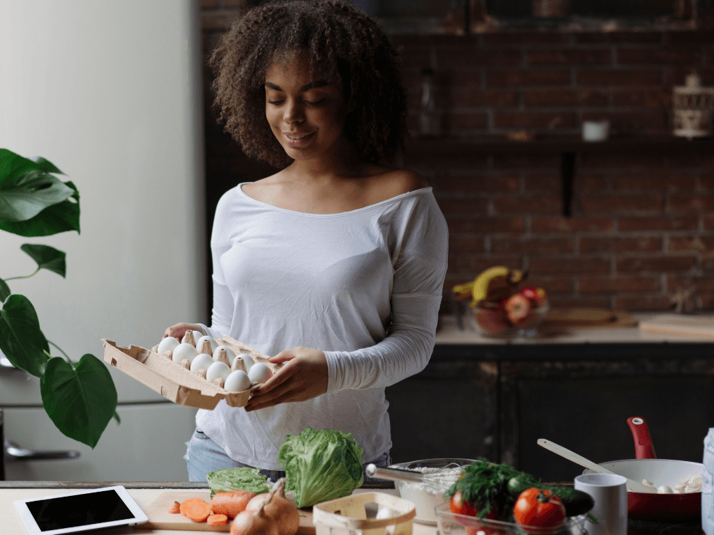A woman in a kitchen with a spread of vegetables, proteins and nutrients meant to nurture the body to help with PMOS