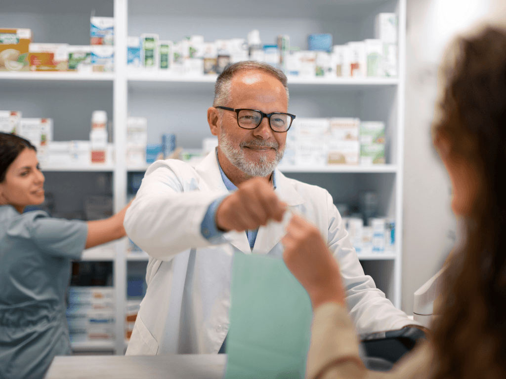 A male pharmacist handing a bag of medication to a woman over the counter, and a female pharmacist in the background reaching for medication on a shelf